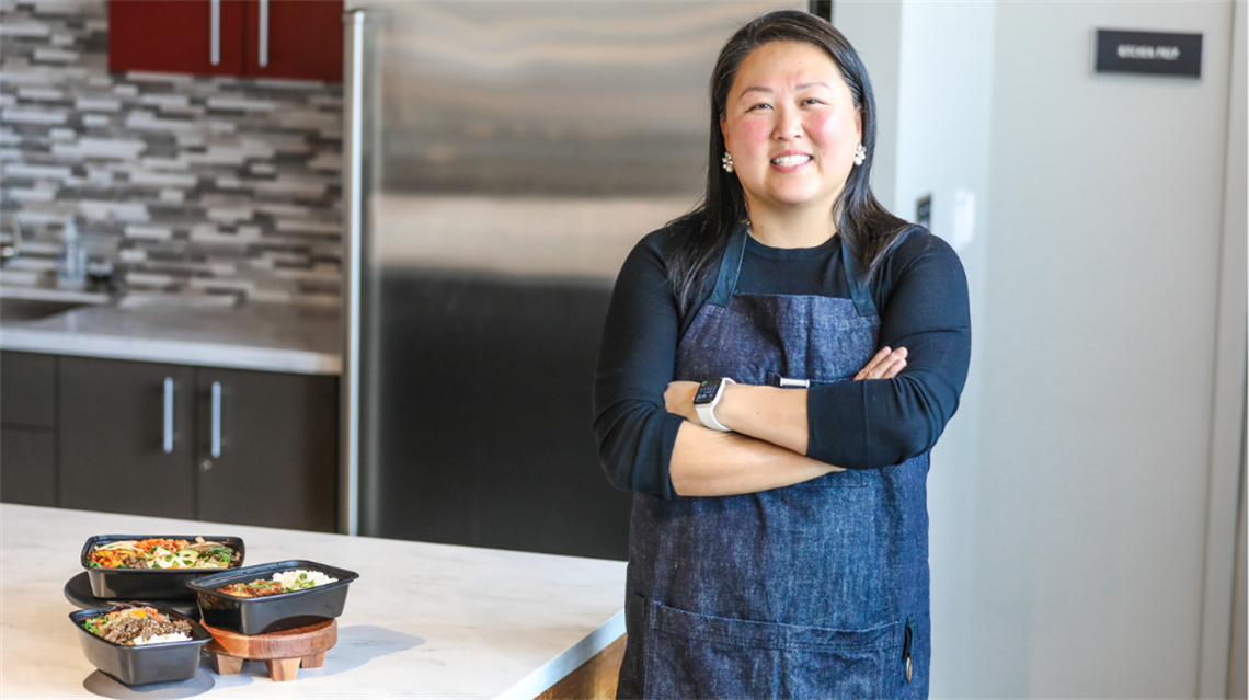 Chef/owner of Kimchi and Bap smiling at camera with three dishes on the counter top.